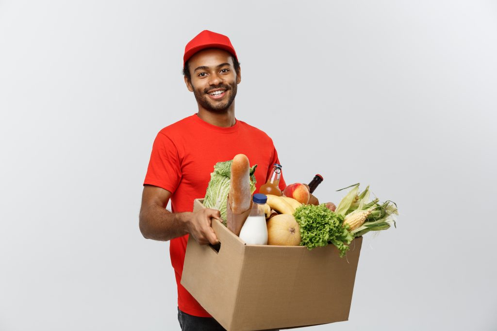 Delivery Concept Handsome African American Delivery Man Carrying Package Box Of Grocery Food And Drink From Store. Isolated On Grey Studio Background. Copy Space. - Simplifiq - Contabilidade para Comércio Varejista e Atacadista em Goiania – GO